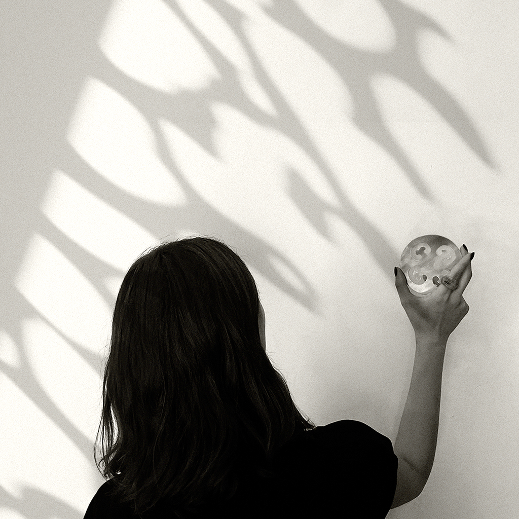lack-and-white photograph of a woman seen from behind, holding a small translucent sculptural object against a sunlit wall. Large organic leaf-like shadows stretch across the wall, creating a quiet interplay of light, nature, and form.