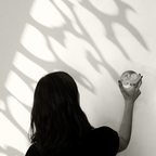 lack-and-white photograph of a woman seen from behind, holding a small translucent sculptural object against a sunlit wall. Large organic leaf-like shadows stretch across the wall, creating a quiet interplay of light, nature, and form.