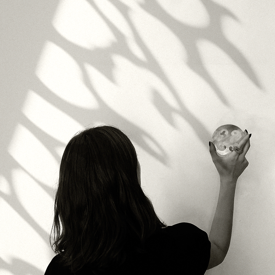 lack-and-white photograph of a woman seen from behind, holding a small translucent sculptural object against a sunlit wall. Large organic leaf-like shadows stretch across the wall, creating a quiet interplay of light, nature, and form.